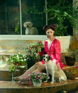 A woman in a red dress poses with dogs and potted plants in a lush garden setting.