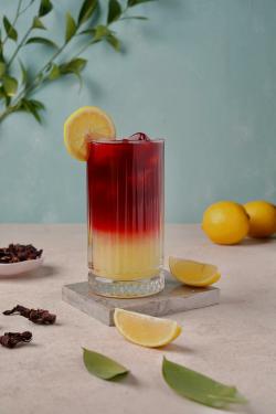 Tall glass of layered lemonade and hibiscus drink garnished with lemon.
