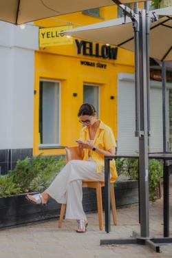 A woman wearing a yellow shirt sits outside a cafe, focusing on her smartphone.