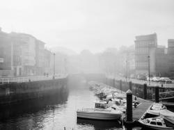Serene black and white image of boats along a foggy riverside with bridge.