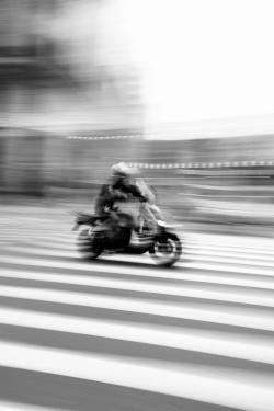 Black and white photo capturing blurred motorcycle crossing a street, illustrating urban motion.