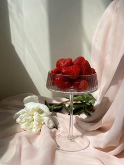 Glass of fresh strawberries on pink fabric with a white flower, bathed in sunlight.