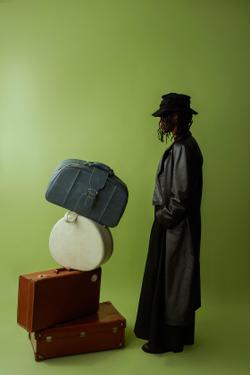 Portrait of a fashionable person in a hat and coat, beside a stack of vintage suitcases.