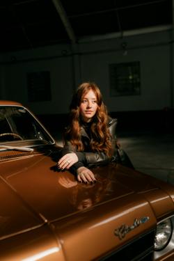 Portrait of a young woman leaning on a vintage car inside a dimly lit garage.