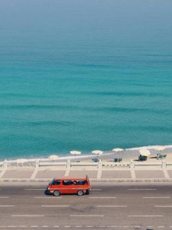 Red van travels along a serene coastal highway with turquoise waters and beach umbrellas.